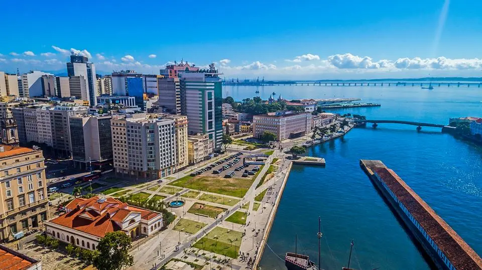 Cidade com vista panorâmica, edifícios variados, parque, rio azul turquesa, ponte e céu claro.