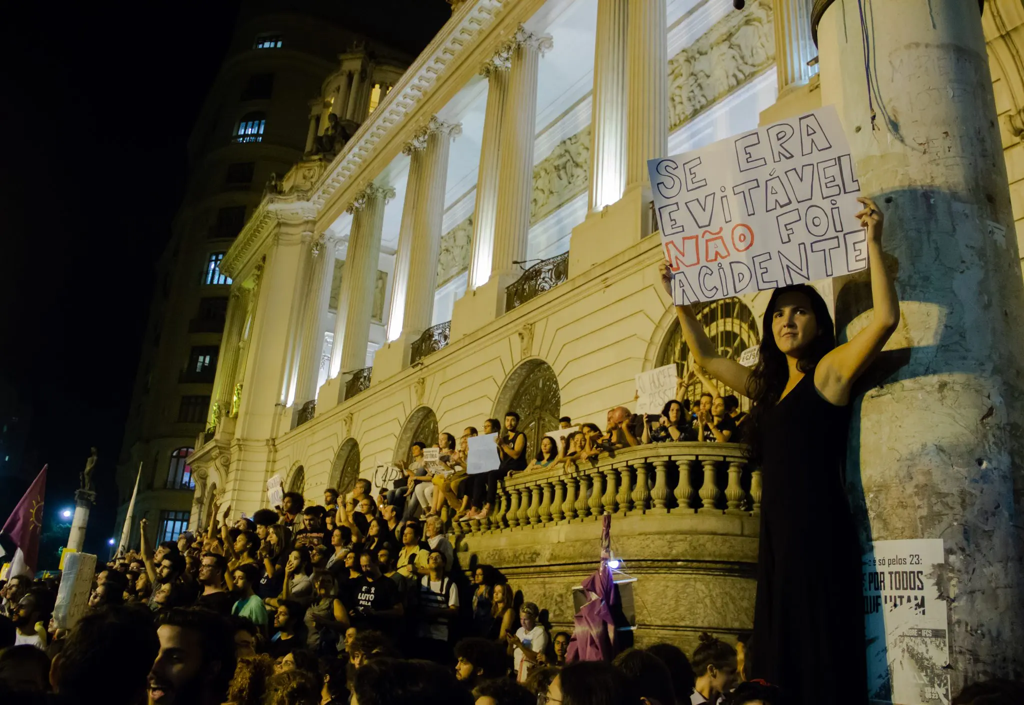 Manifestação noturna em frente a edifício neoclássico.