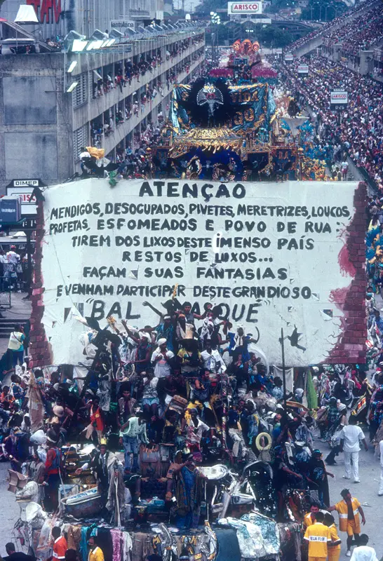 Desfile de Carnaval com carro alegórico e cartaz sobre grupos sociais.