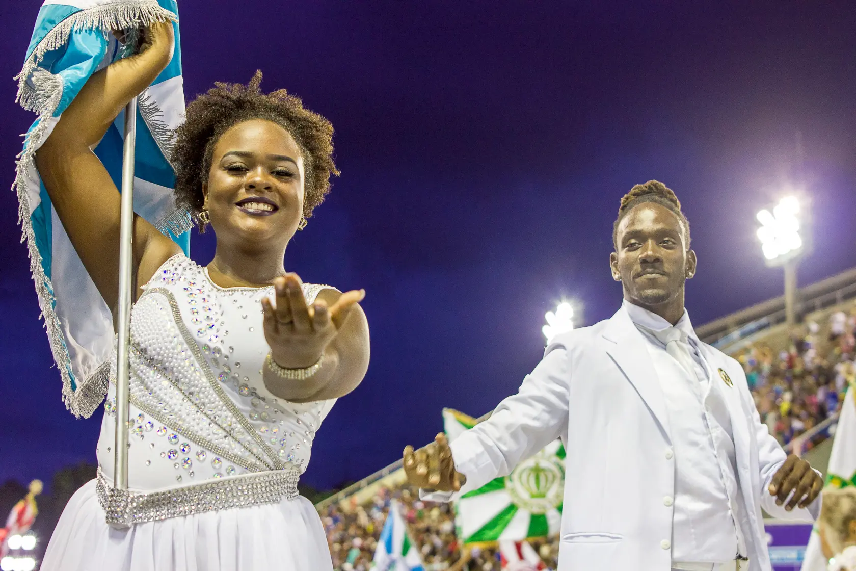 Fotografia de desfile noturno com mulher em traje branco e bandeira azul, sorrindo e fazendo gesto.
