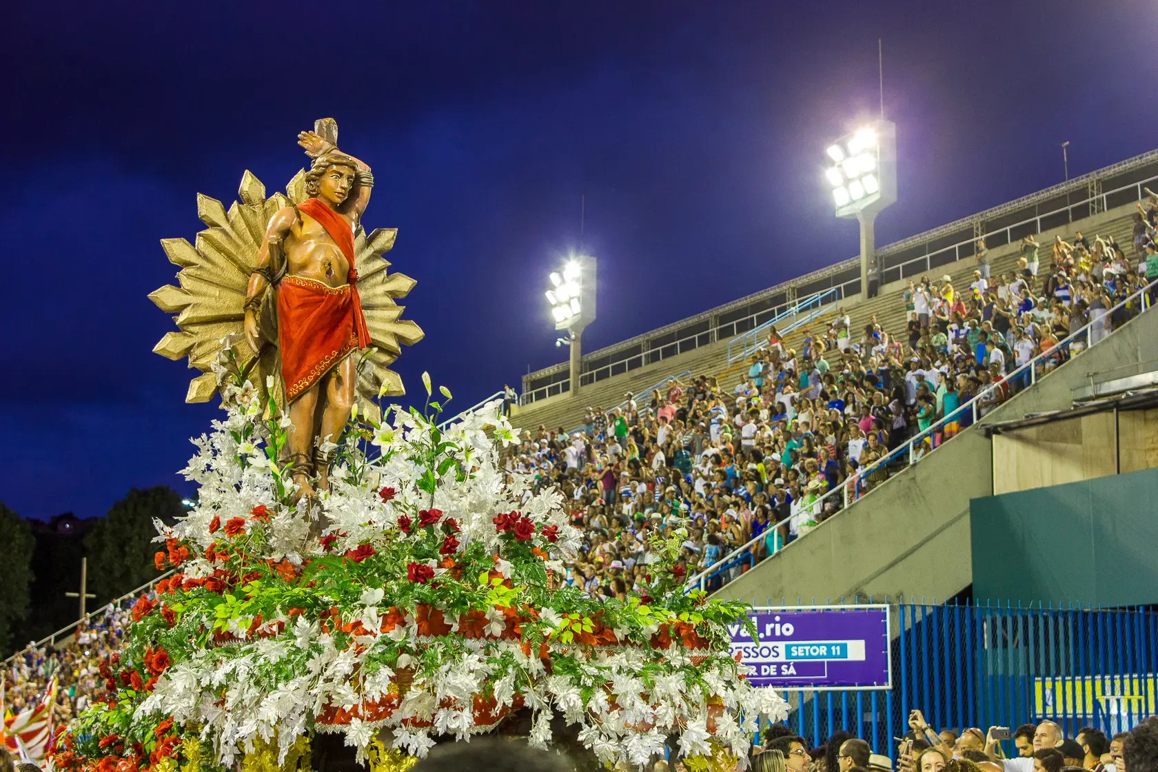 Fotografia de procissão noturna com estátua central e multidão assistindo.