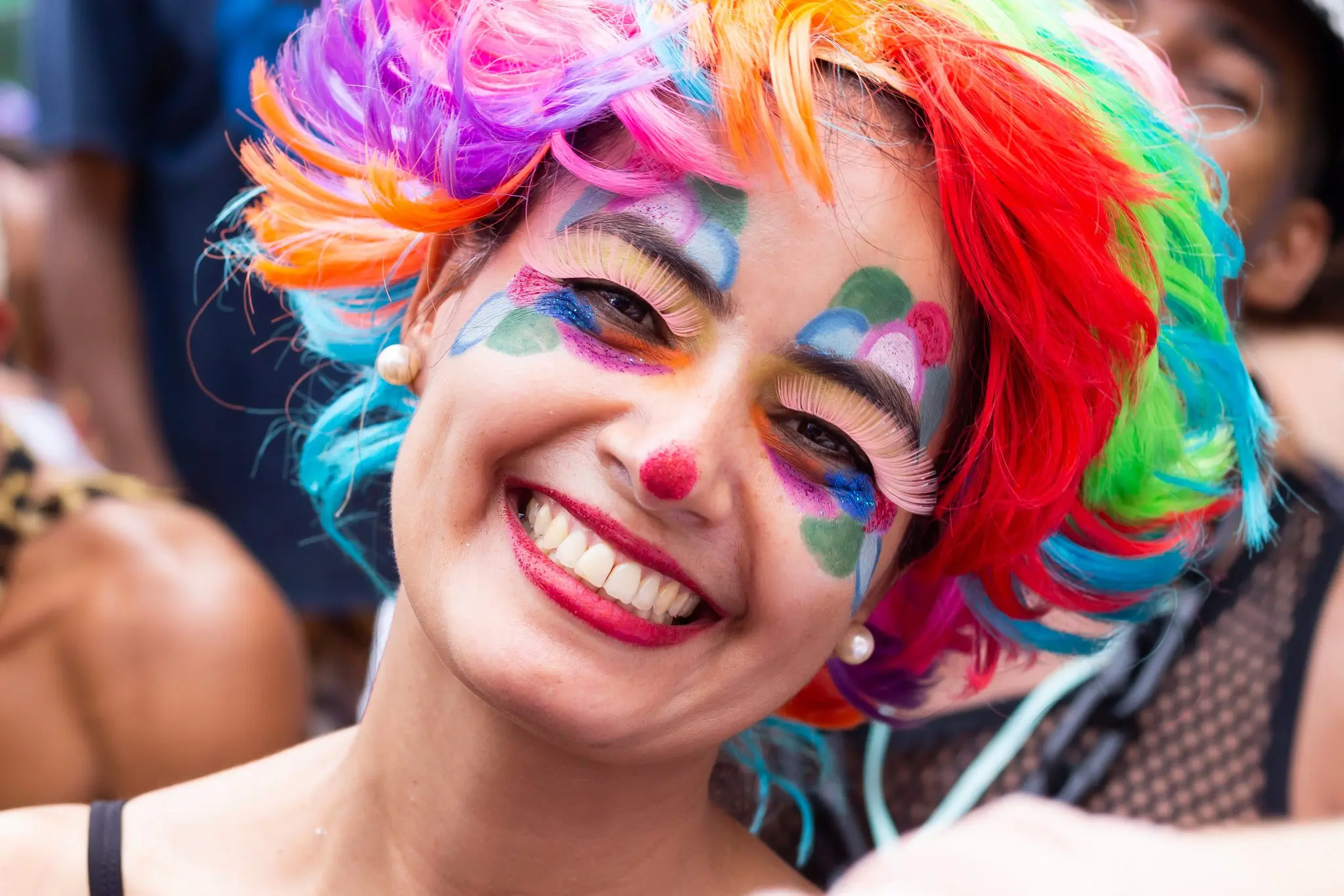 Pessoa sorridente com penteado colorido e rosto pintado, ouvidos com pérolas.