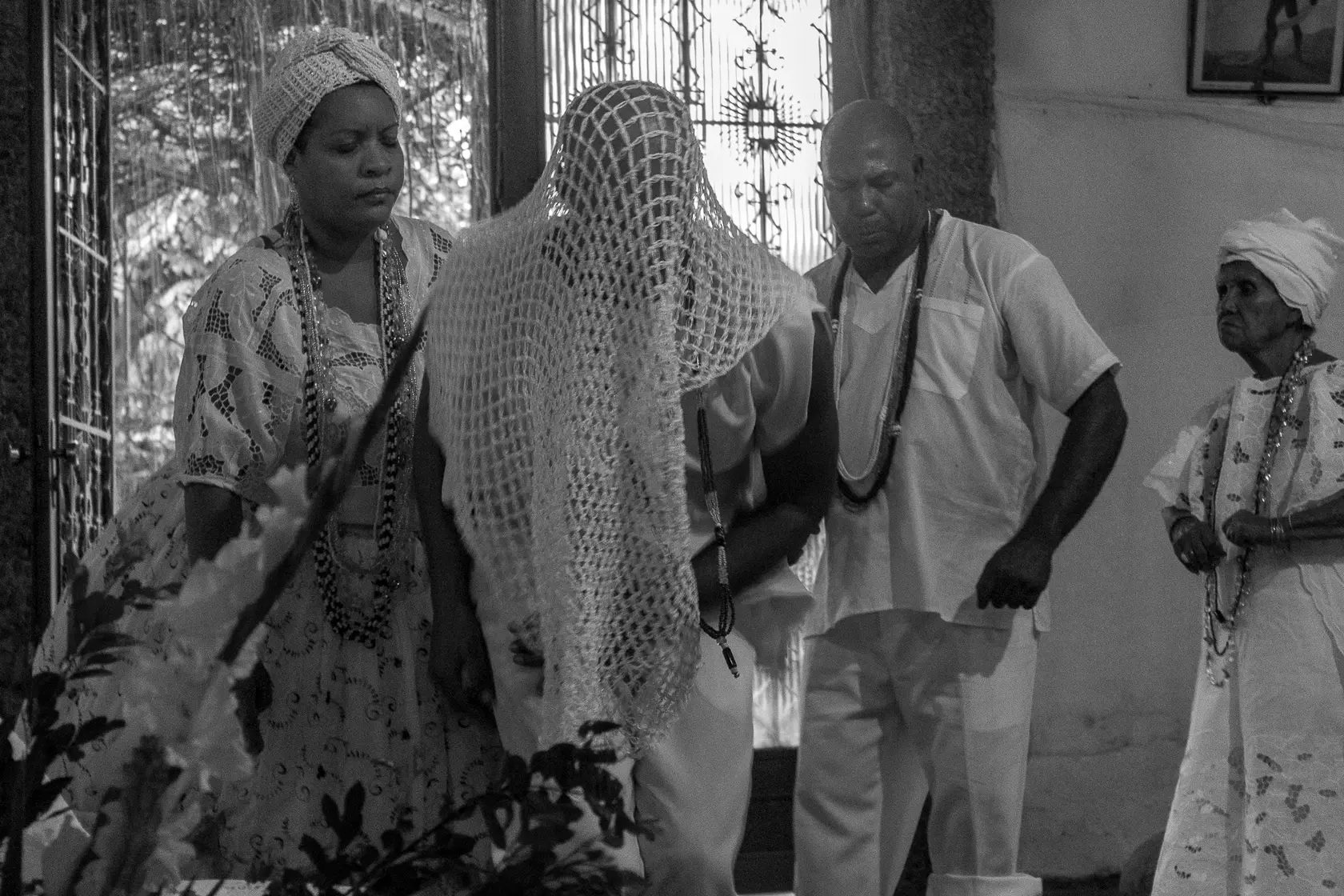 Mulher com vestido bordado segura vara de bambu; figura coberta por rede; homem de pé com colar; mulher mais idosa olha figura coberta; janela com grades e imagem religiosa.