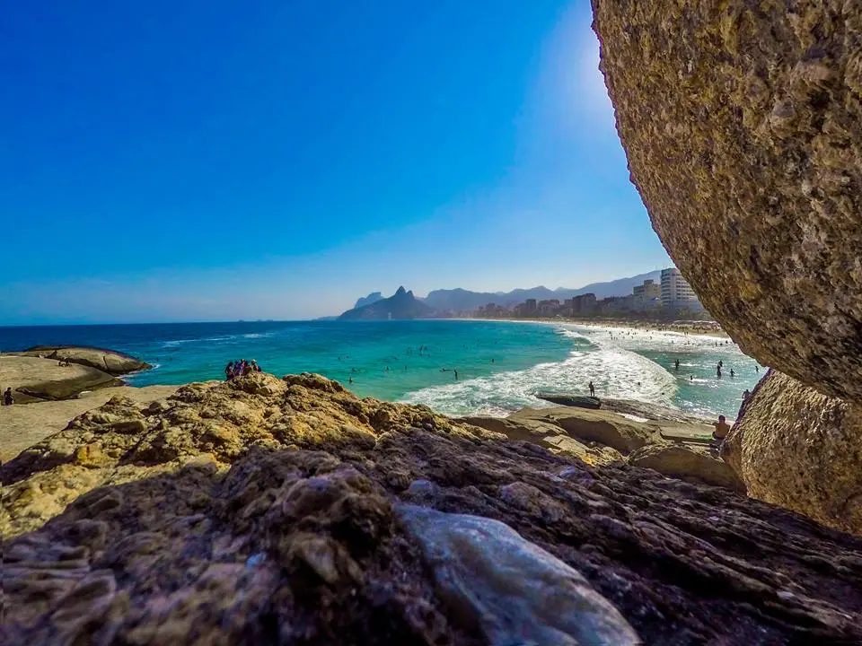 Praia com céu azul, rochas arenosas, mar turquesa, edifícios e montanhas.