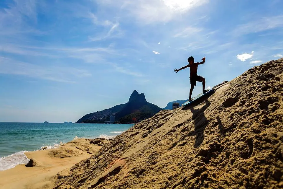 Menino equilibra-se na praia com colina de areia, montanha rochosa e edifício moderno ao fundo.