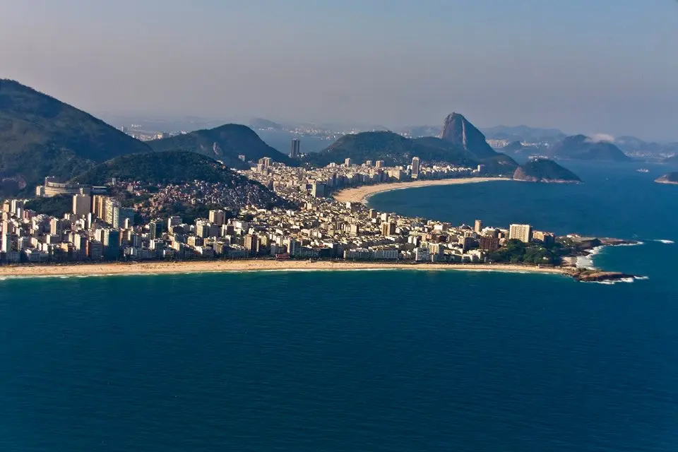 Paisagem costeira com montanhas, cidade, praia e mar calmo.