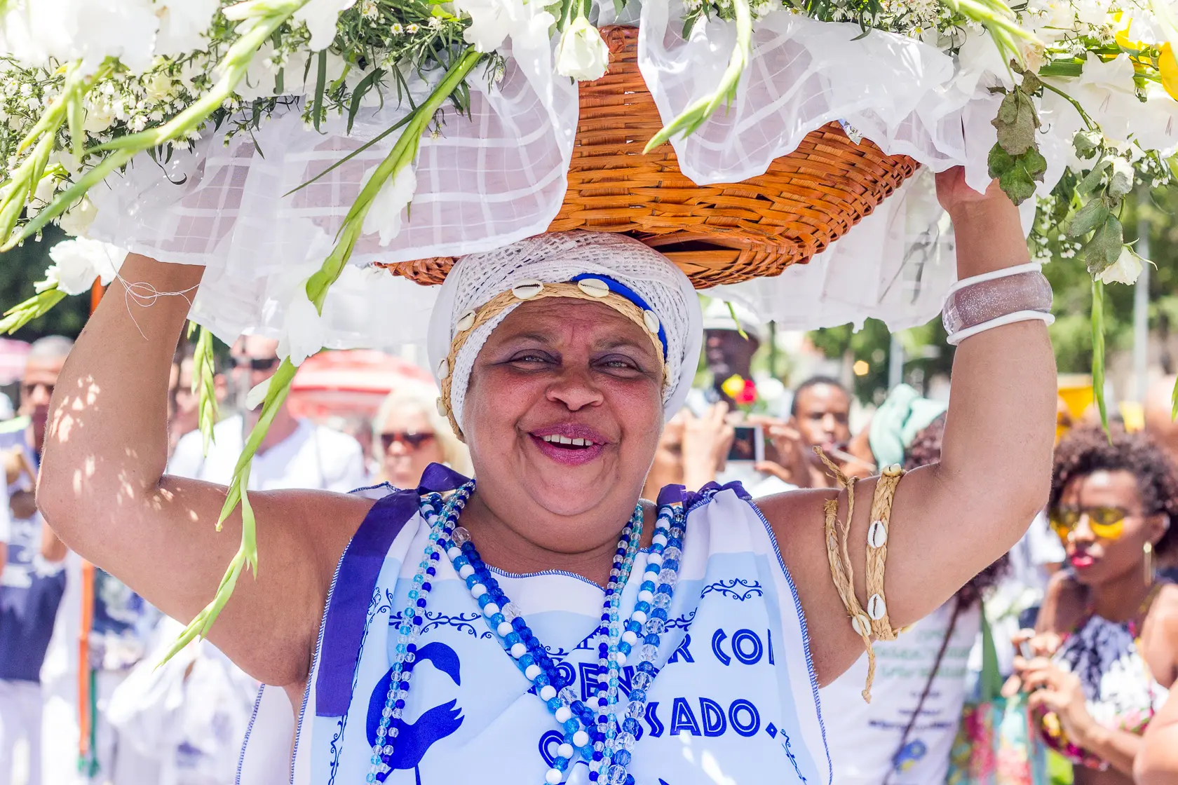 Mulher sorridente com traje tradicional azul e branco, segurando cesto de flores.