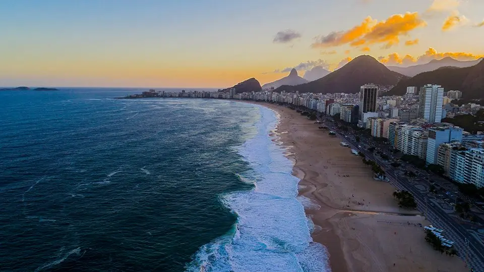 Praia ao pôr-do-sol com oceano, cidade e montanhas.