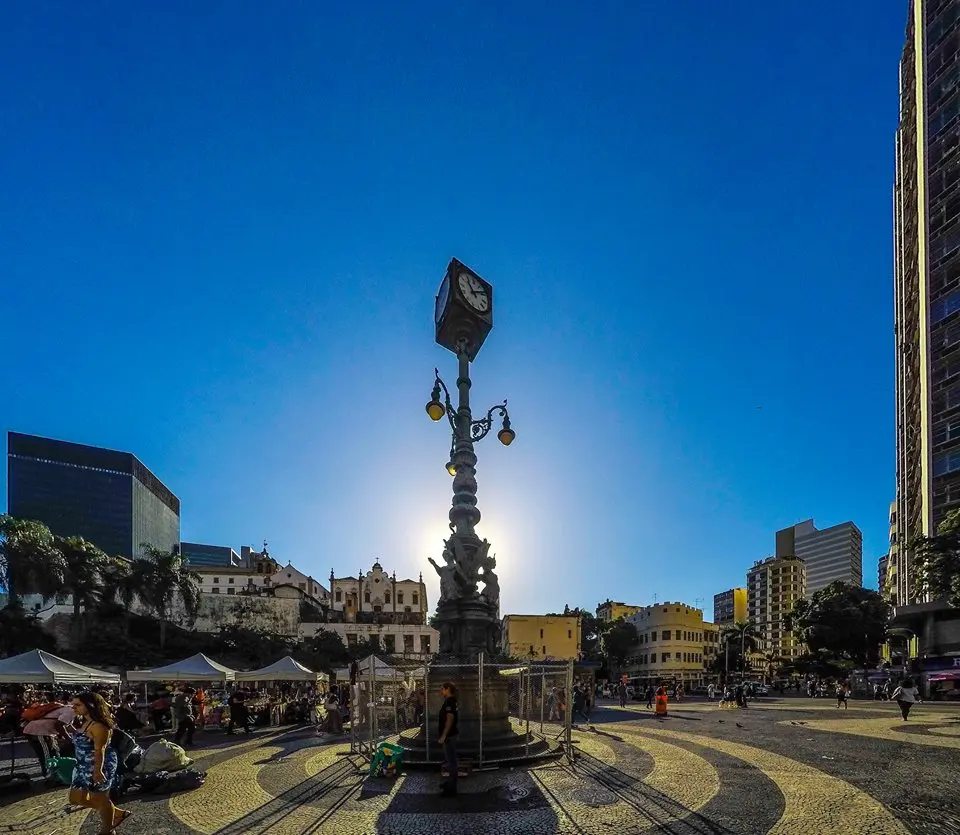 Praça com relógio ornamentado sobre coluna, feira de rua e casario histórico ao fundo, sob céu azul.
