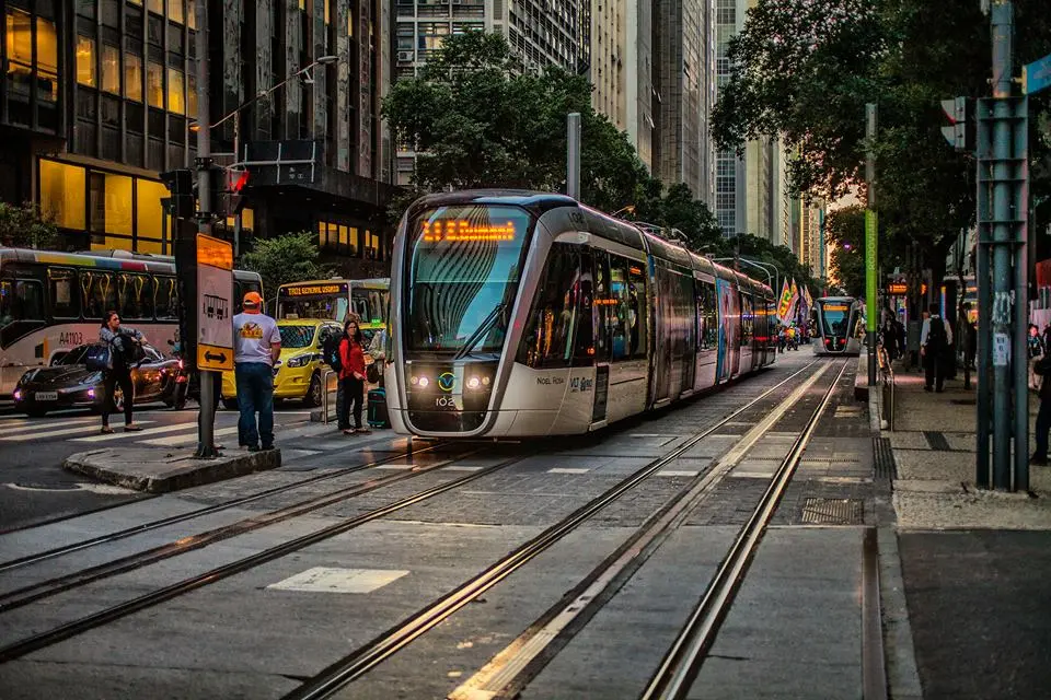 Uma rua urbana com ônibus, trens e pessoas em movimento.