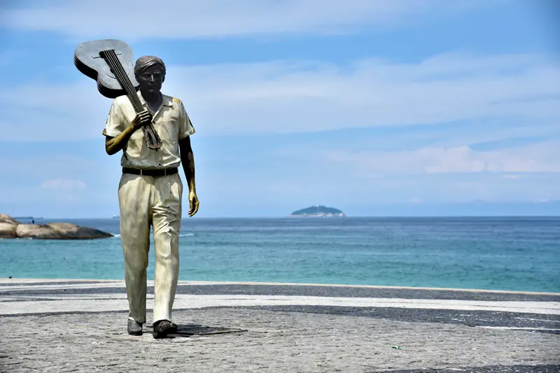 Uma estátua de homem com guitarra na praia, mar e ilha ao fundo.