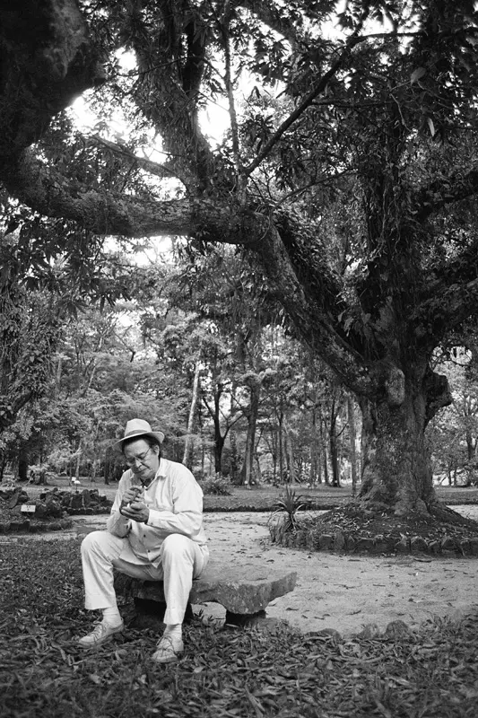 Homem em banco de pedra no Jardim Botânico do Rio, vestindo chapéu e olhando para baixo.