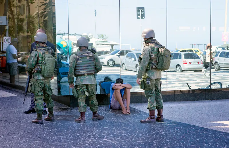 Três fuzileiros navais em uniforme militar observam dois homens submissos em um ambiente urbano.