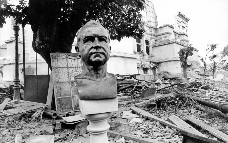 Busto de Getúlio Vargas em frente a edifício em ruínas.