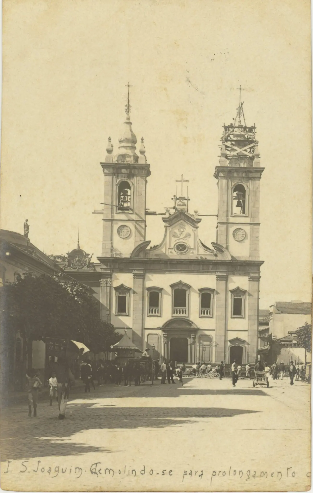 Igreja antiga com torres laterais e frontispício adornado, povoada por pessoas e carruagens em praça, céu claro.