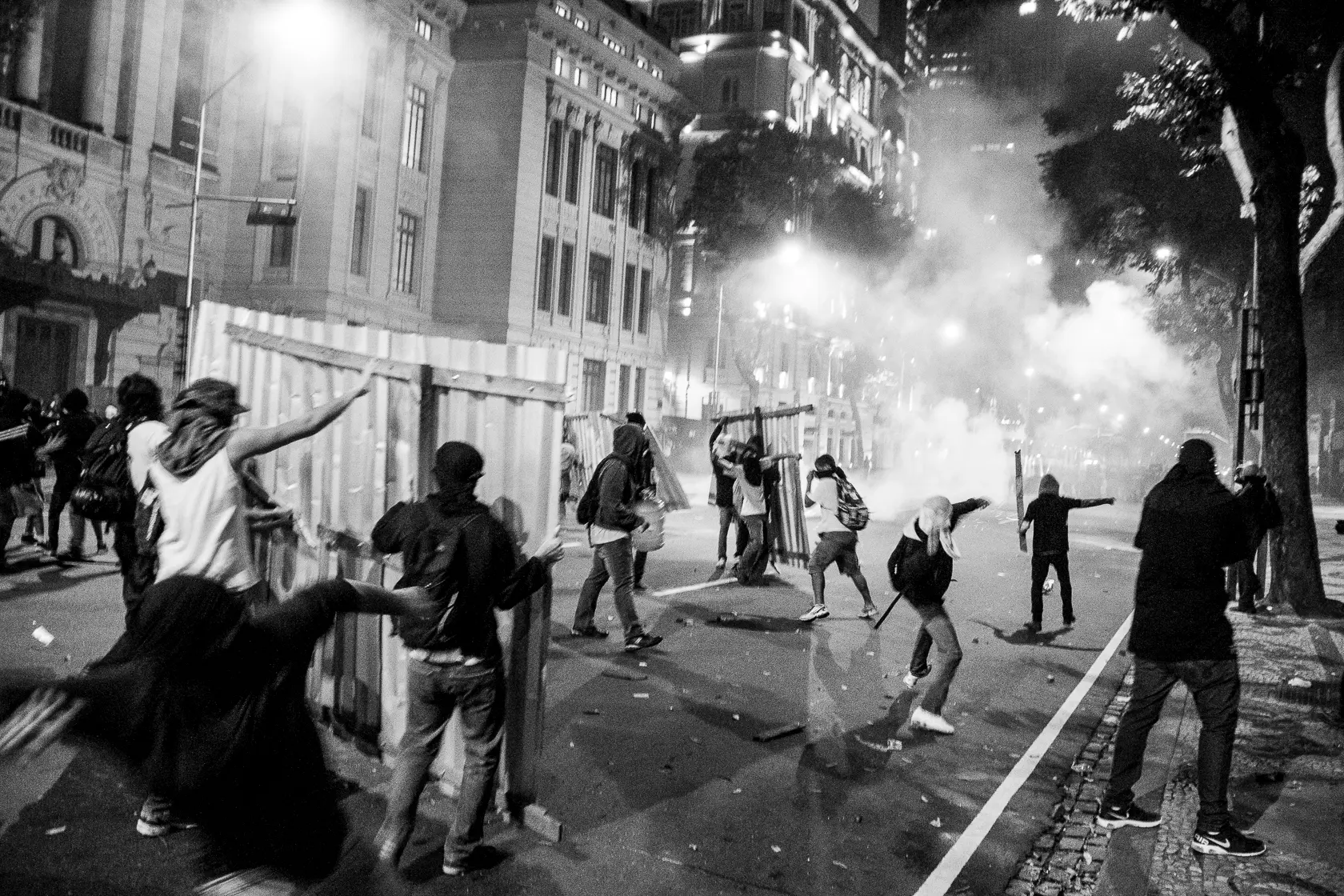 Cena noturna urbana com pessoas em movimento e fumaça sugerindo confrontos.