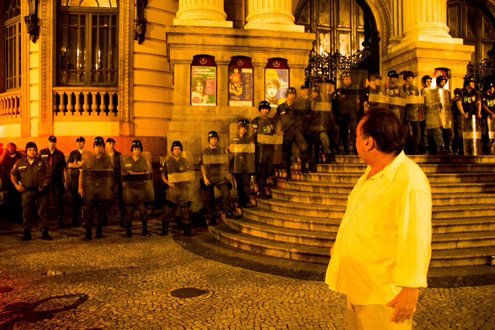 Cena noturna com policiais em fila segurando escudos, homem de costas olhando-os, edifício monumental com quadros decorativos.