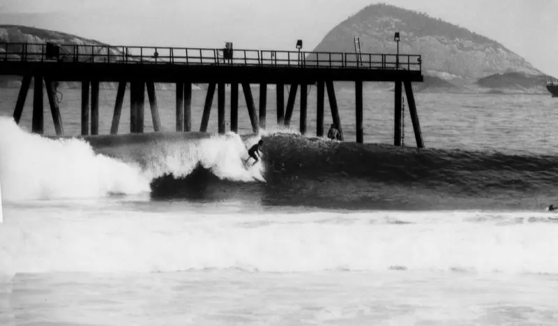 Praia com píer e surfista em onda.