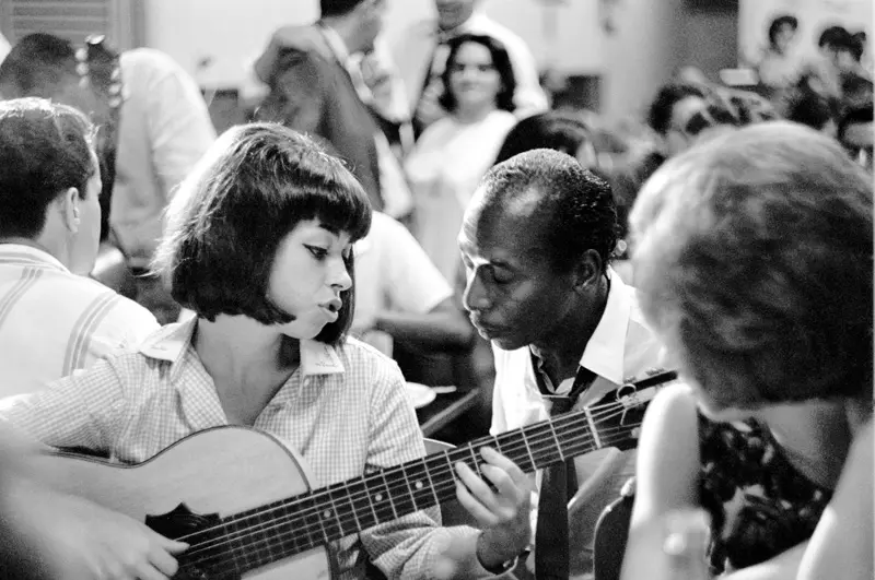 Uma mulher toca violão enquanto um homem assiste em um salão social.