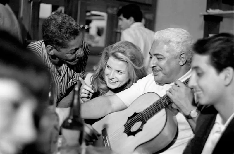 Quatro pessoas sorridentes em um ambiente social, dois homens e uma mulher com guitarra.