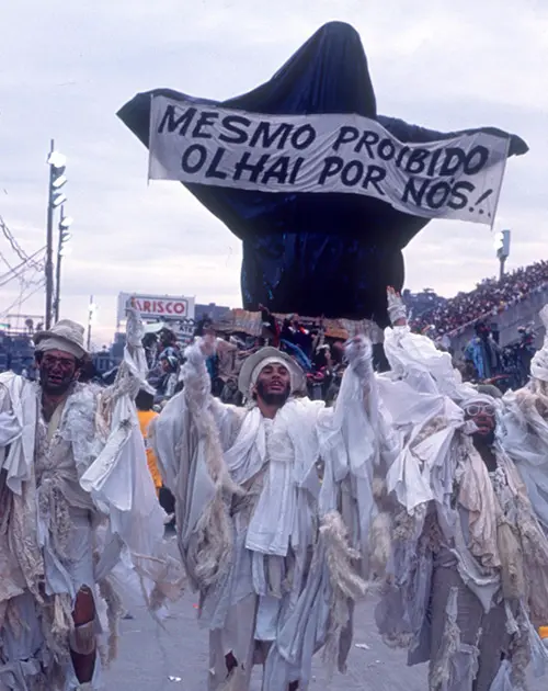 Desfile de escola de samba em 1989, figura com capa preta segurando cartaz.