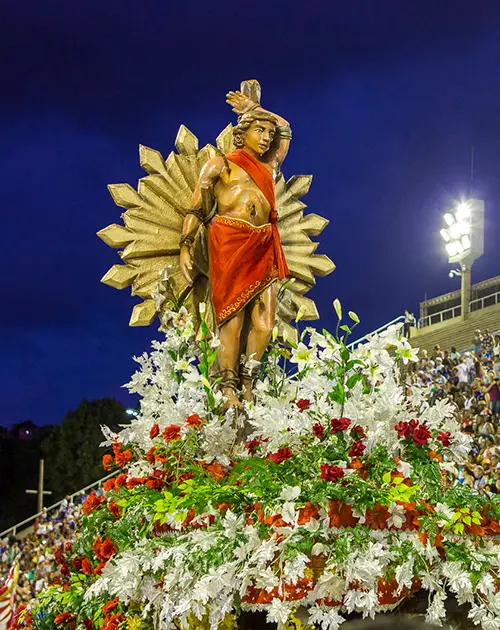 Desfile de Carnaval com estátua masculina adornada com flores e coroa dourada.