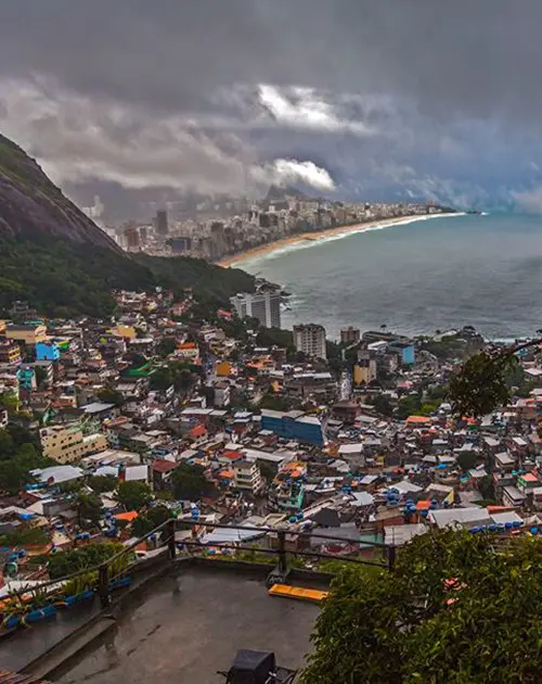 Cidade costeira sob céu nublado com montanha verde, edifícios coloridos, mar com ondas e prédios modernos ao fundo.