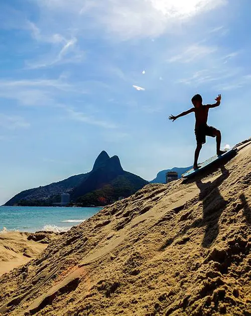 Menino equilibra-se em prancha de surf na praia, com oceano e montanha ao fundo.