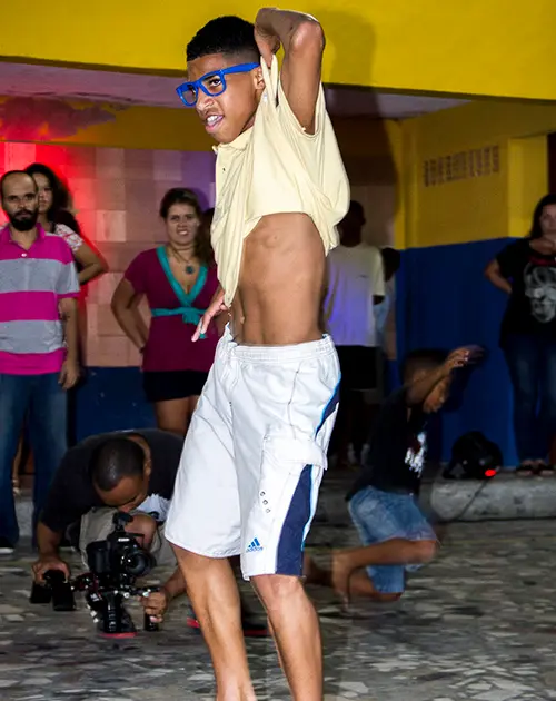 Homem dançando em evento festivo, com decoração colorida ao fundo.