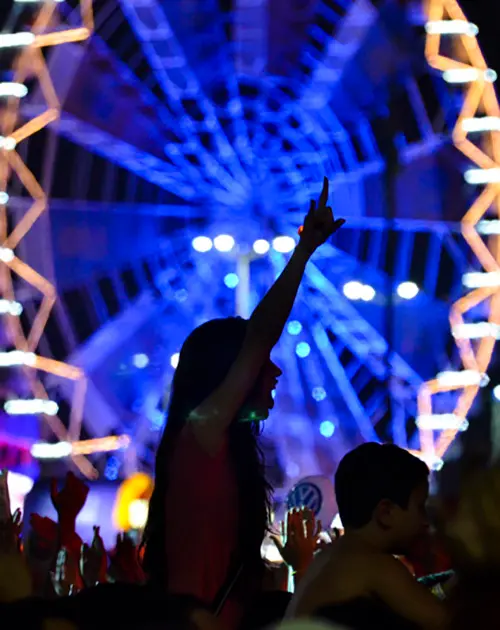Fotografia de evento noturno com roda-gigante iluminada, foco em mulher com cabelos longos vestindo vermelho levantando o braço.