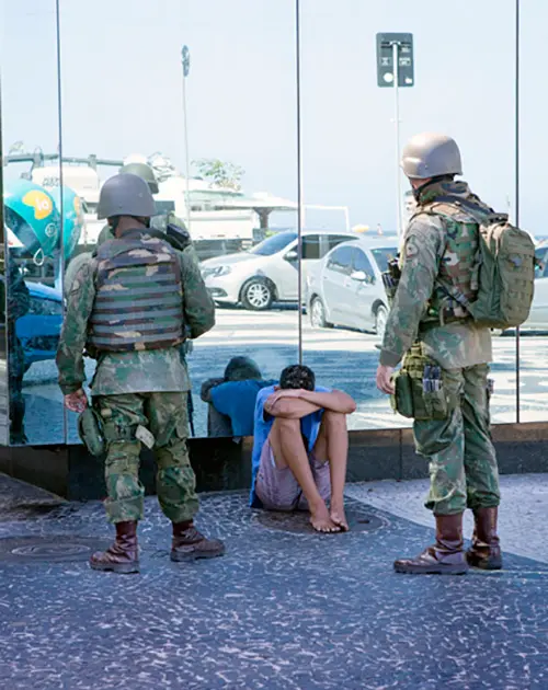 Três soldados em uniforme observam figura sentada no chão em cenário urbano.