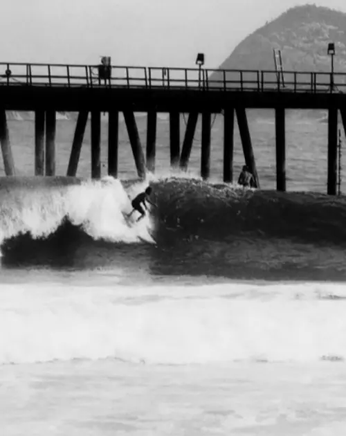Surfista em ação na praia com píer ao fundo.