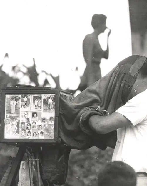 Homem com câmera vintage, revelando fotos impressas; figura de pessoa de pé ao fundo.