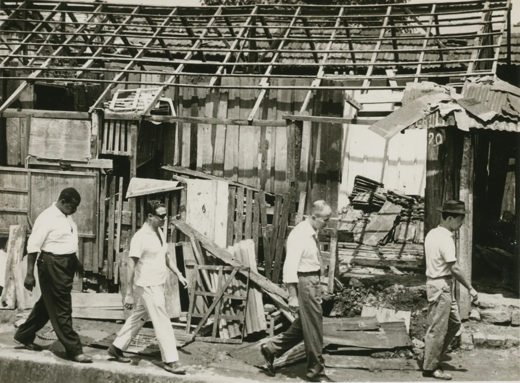 Fotografia preto e branco de favela em remoção, com três homens e construções em destaque.