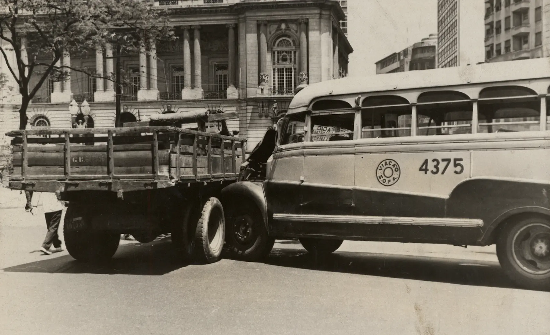 Caminhão de carga com plataforma aberta, pessoa caminhando à frente, ônibus antigo parado na rua.