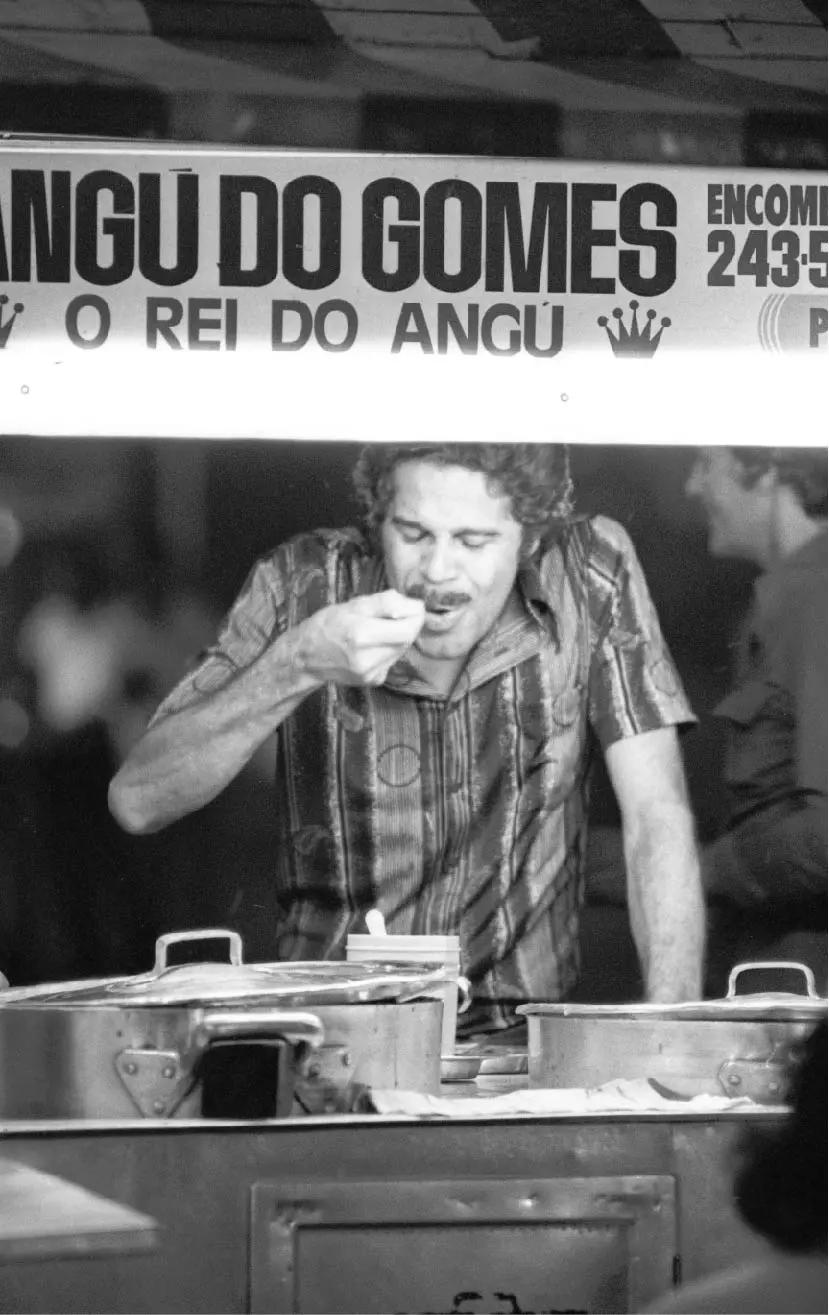 Homem degustando em cozinha com panelas e banner mencionando "Angú do Gomes".