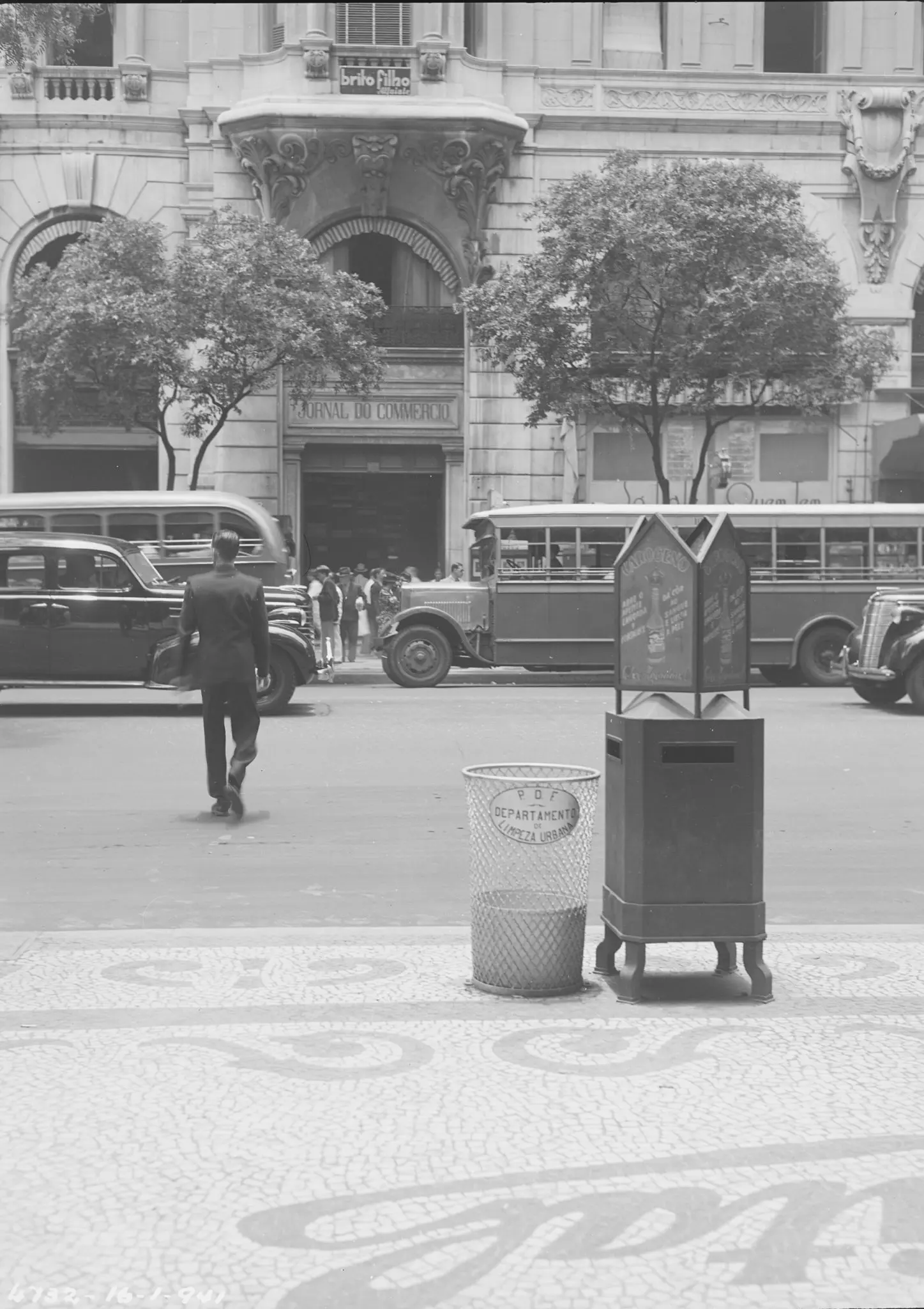 Fotografia preto e branco de rua urbana com lixeiro, dispositivo de coleta, homem em terno, veículos antigos e prédio clássico.