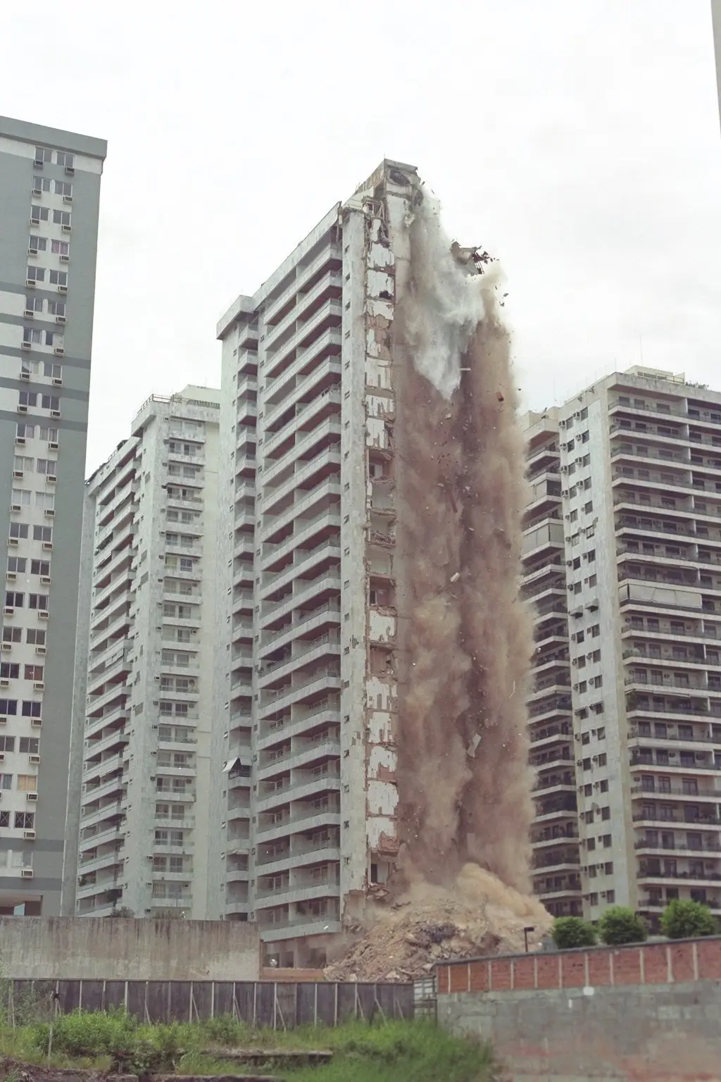 Desabamento de edifício com prédios cinzentos e nuvem de poeira.