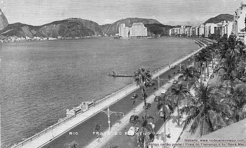 Praia com avenida paralela à água, mar e montanhas ao fundo.