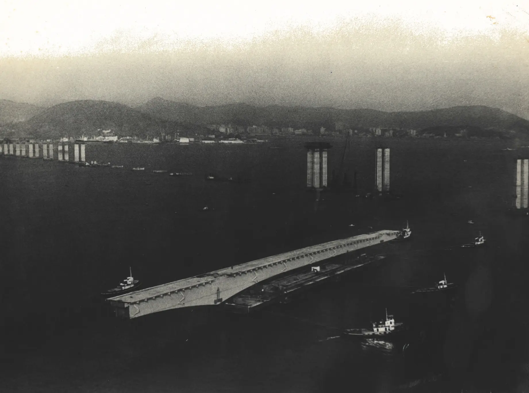 Ponte sobre rio com barcos e montanhas ao fundo.