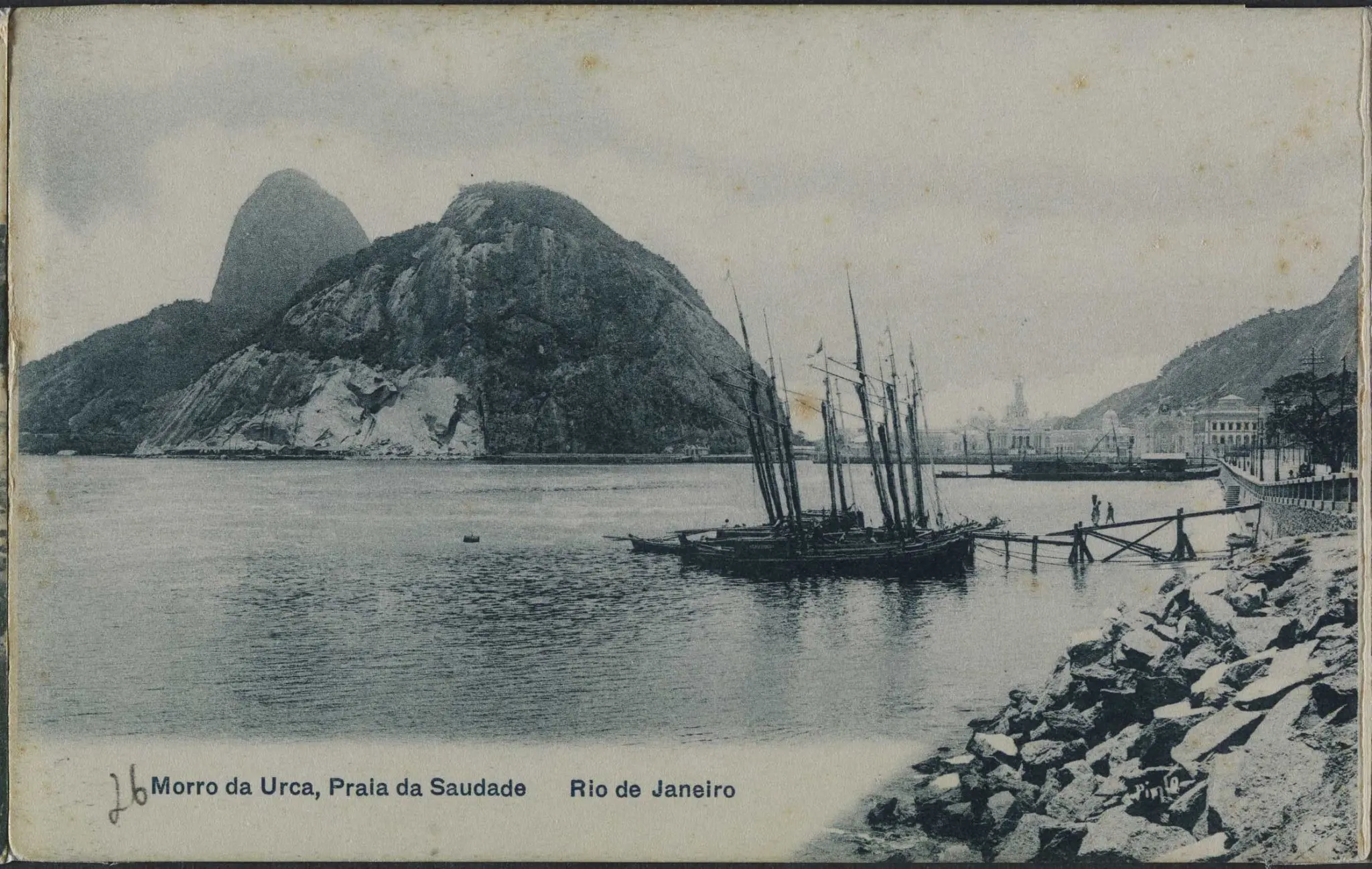 Praia com montanhas, rochedos, edifícios, navio a vela, pedras e ponte de madeira.