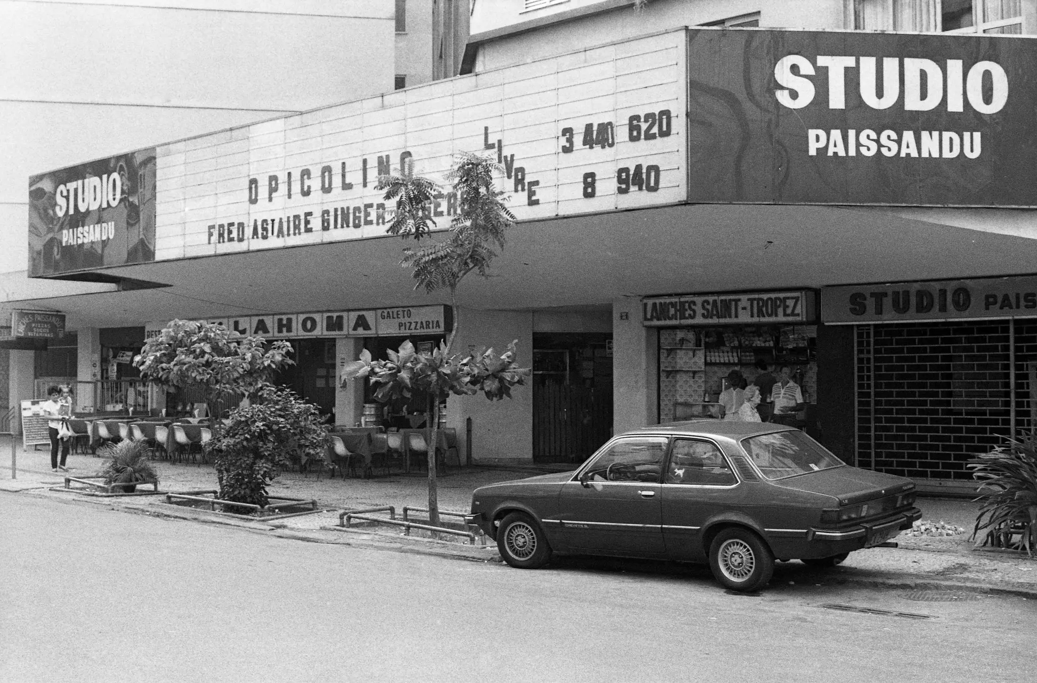 Fachada preto e branco de cinema "Studio Paissandu" com estacionamento, árvores, lojas e filmes anunciados.