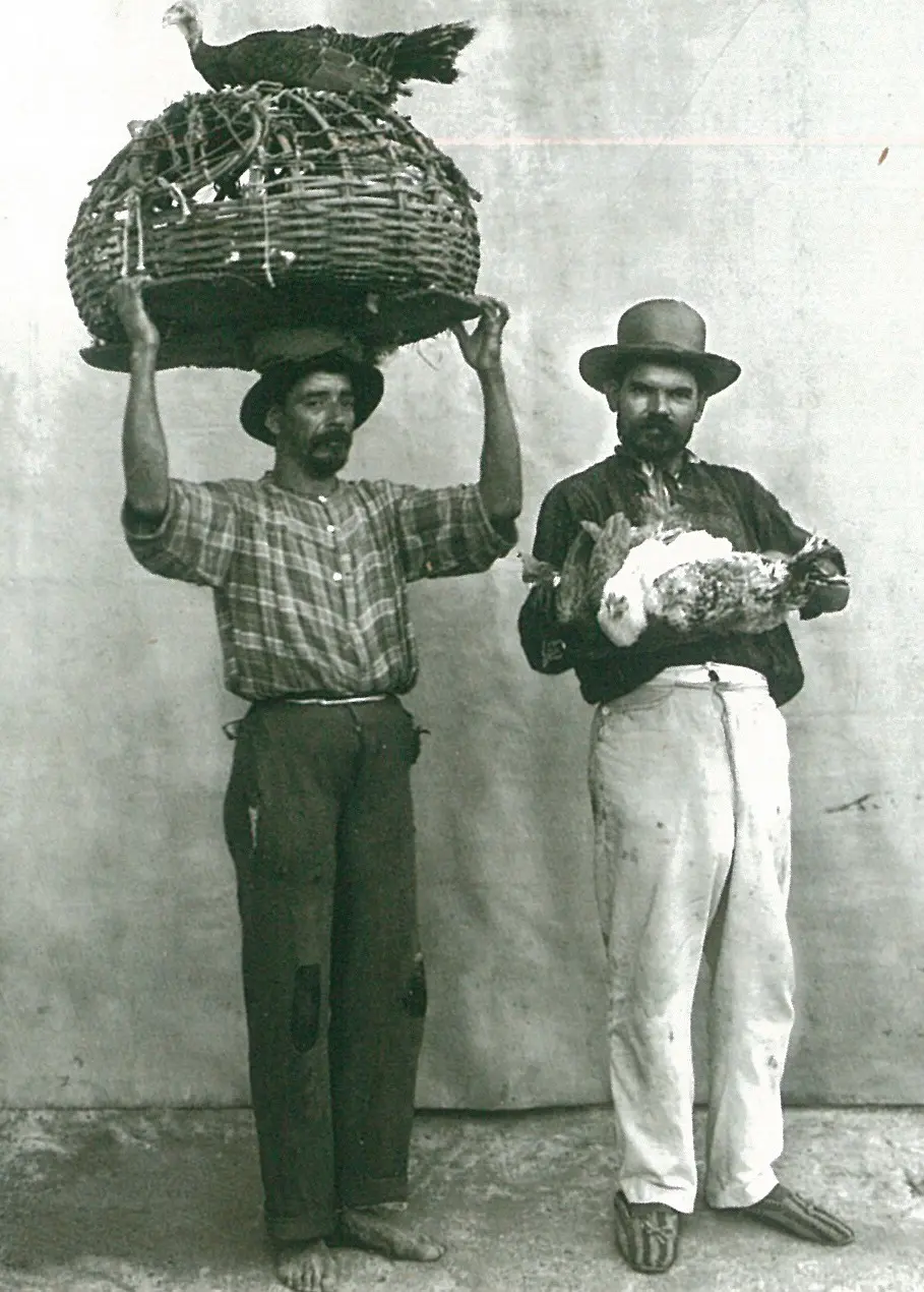 Dois homens em preto e branco, um segurando uma cesta com uma ave e o outro um animal vivo.