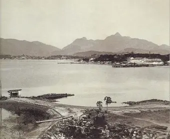 Praia arenosa com montanhas ao fundo, prédios e árvores.