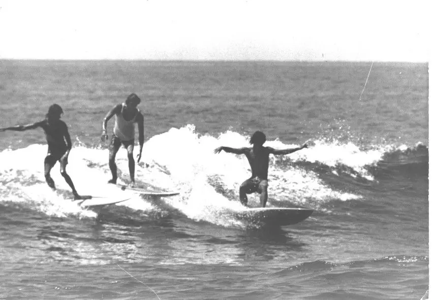 Três pessoas praticando surf na praia, em preto e branco.
