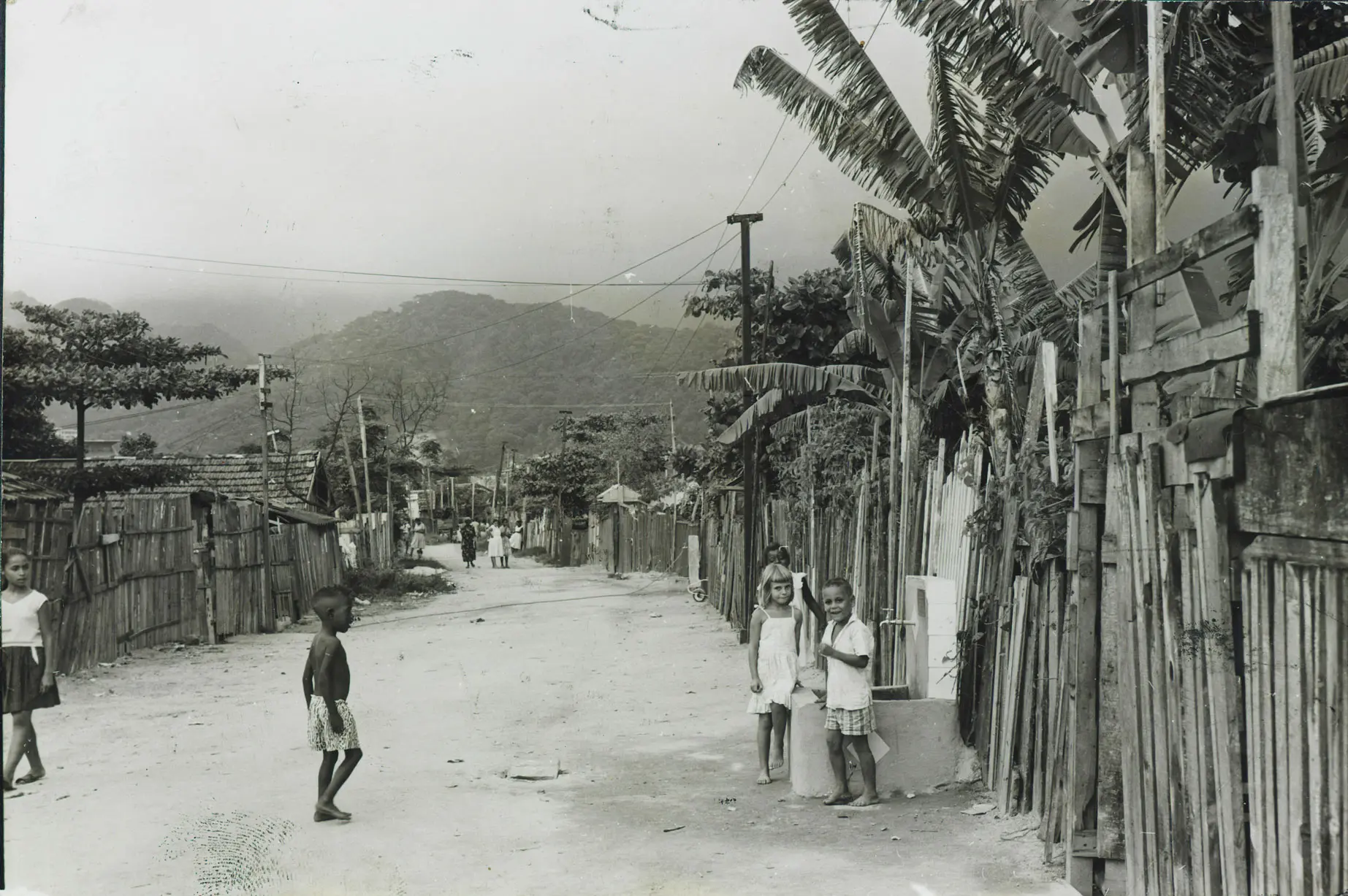 Favela em preto e branco, ruas estreitas, casas simples, bambu, crianças caminhando, colina verde, cabos elétricos, árvores folhosas.
