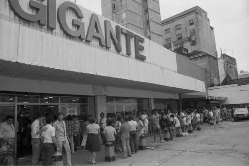 Fila longa em supermercado "Disco Gigante" com prédios urbanos ao fundo.