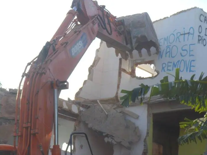 Escavadeira demolindo edifício branco com céu azul.