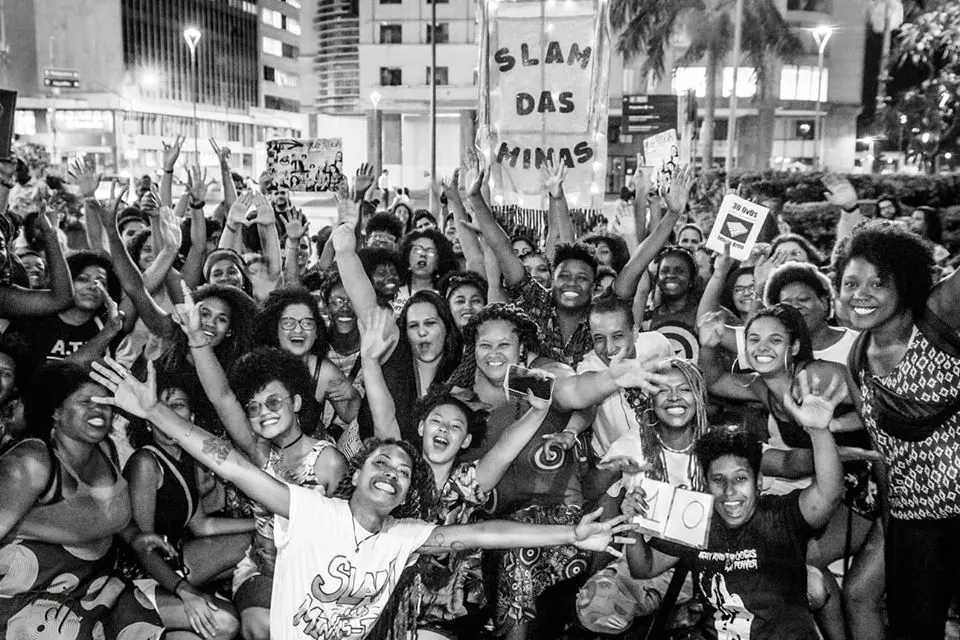 Multidão animada com braços levantados, celular e cartaz "SLAM DAS MINAS" em fundo urbano noturno.