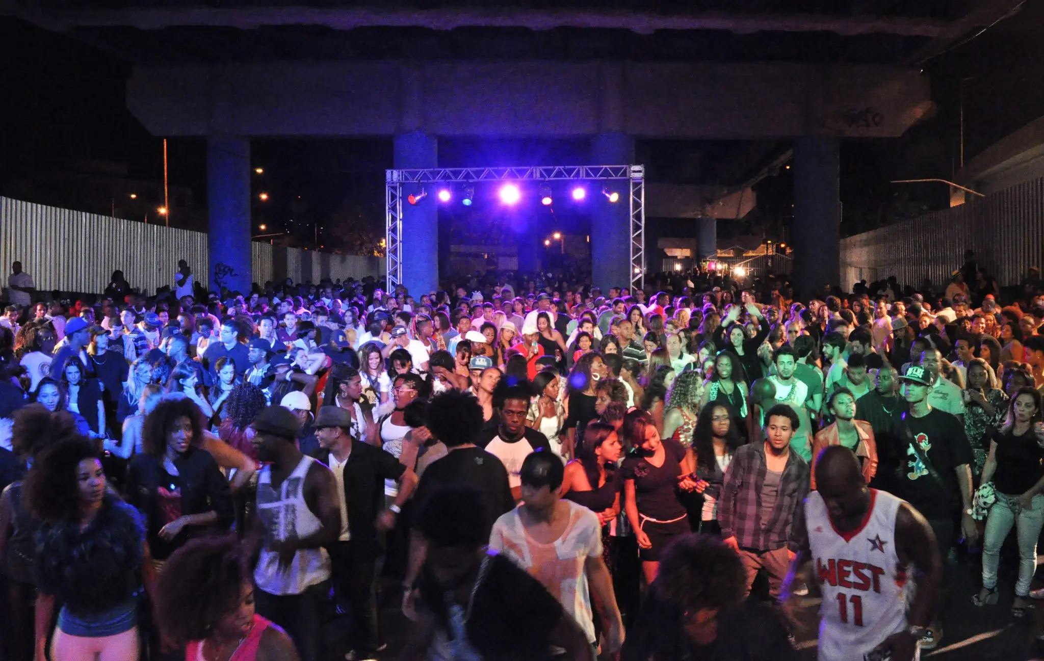 Multidão dançando em evento festivo sob ponte, com palco iluminado.